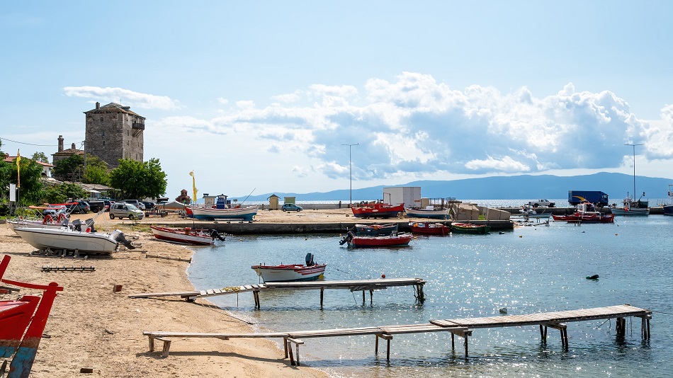 Sea port, moored boats on Aegean sea, few parked cars, two small wooden piers, hills on the background and Tower of Prosphorion, Ouranoupolis, Greece